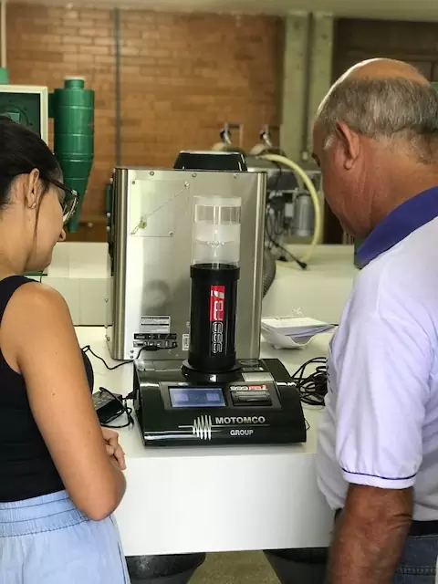 Uma jovem mulher e um homem mais velho estão olhando atentamente para um equipamento de medição da marca Motomco. Eles estão em um ambiente que parece ser um laboratório ou uma oficina. O equipamento possui uma tela digital e está ligado, indicando que está em uso. A mulher está à esquerda, com cabelo preso e vestindo uma blusa preta, enquanto o homem está à direita, com cabelo grisalho e vestindo uma camisa branca com detalhes em azul.